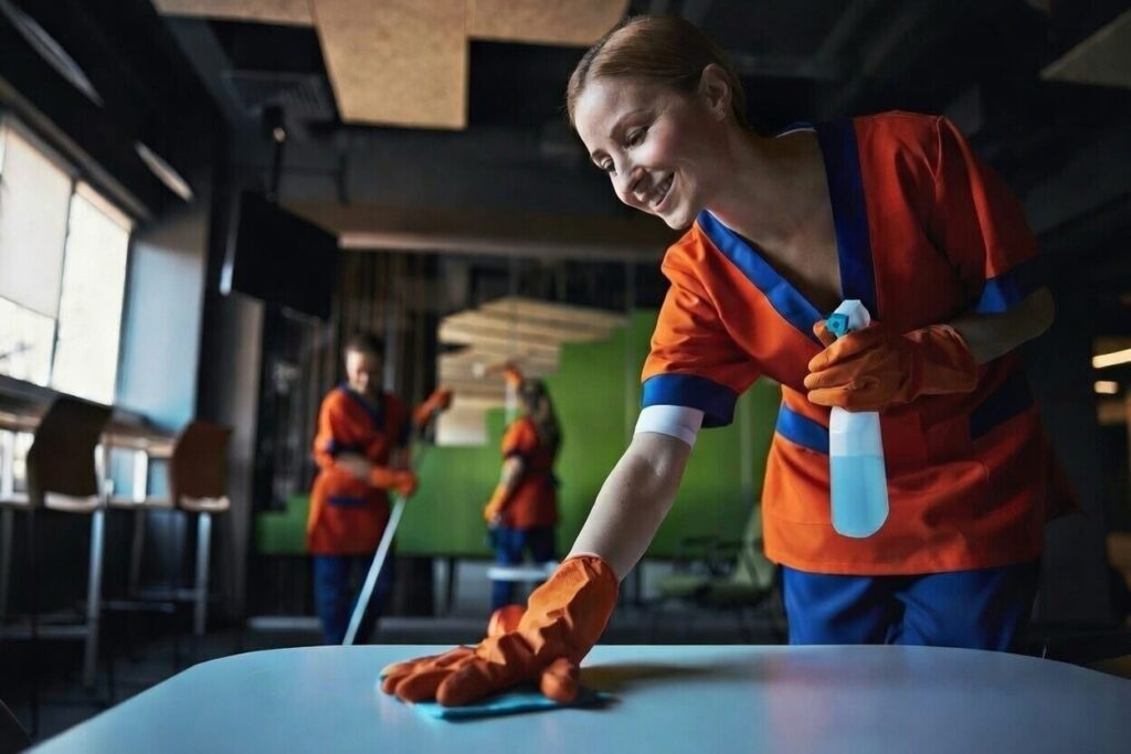 A woman in orange cleaning uniform and gloves wipes a table with a cloth and spray bottle, smiling, while two coworkers clean in the background of a modern, well-lit room.