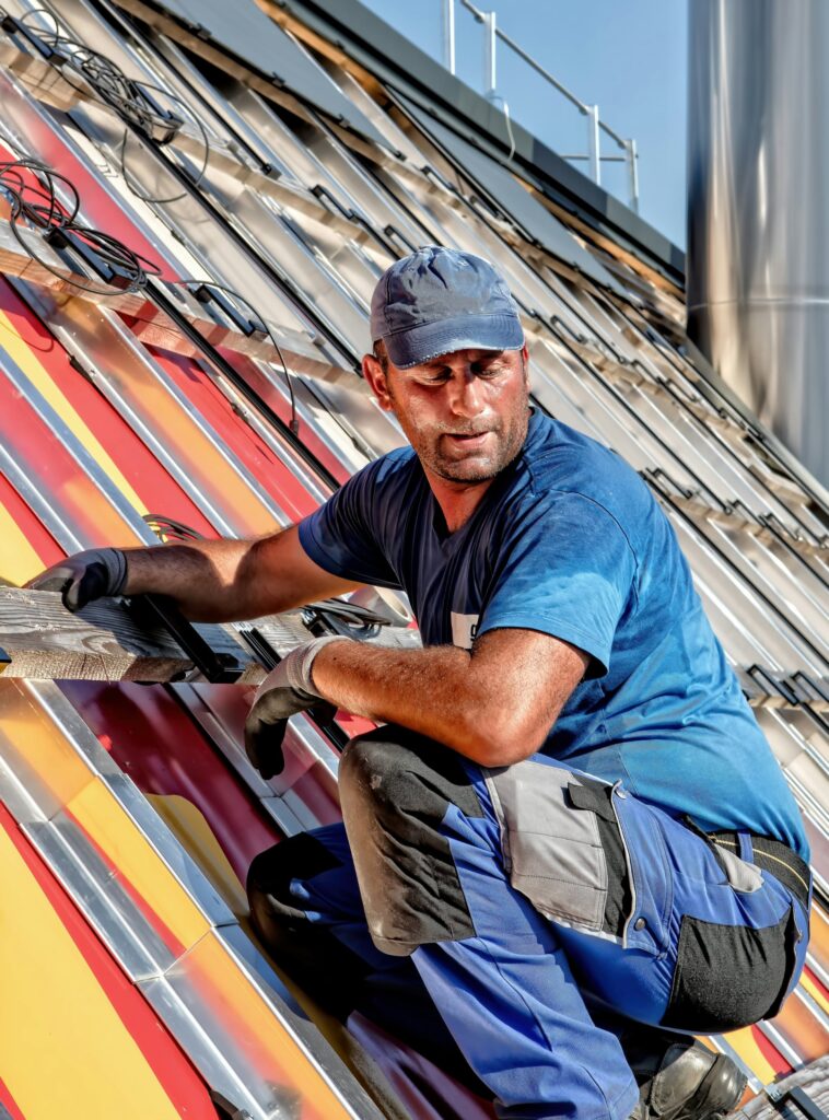 A construction worker in blue workwear and a cap installs metal panels on a sloped roof, holding a tool and wearing gloves, under bright sunlight.
