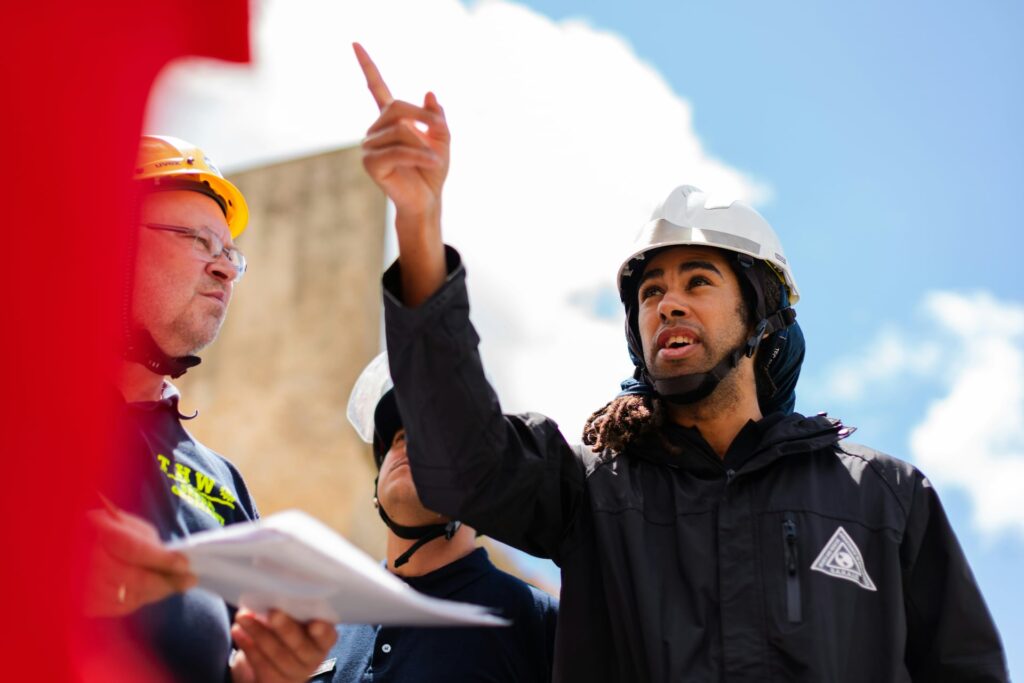 A man in a safety helmet and jacket points upward while speaking to two colleagues, one holding papers, at an outdoor construction or work site under a bright sky.