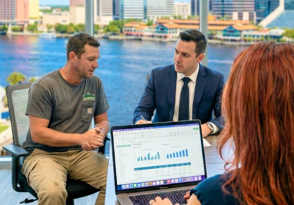 Three people meet in a modern office with city views; one in a suit speaks to a casually dressed man, while a woman with a laptop showing graphs sits at the table.